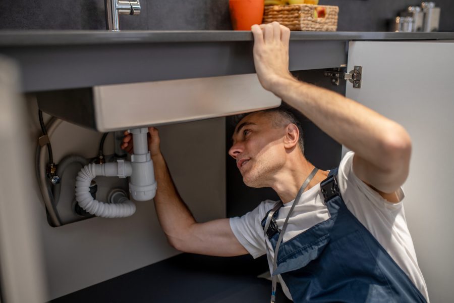 Man carefully examining bottom of sink and pipe Plumber, repair. Experienced attentive middle-aged man carefully examining bottom of kitchen sink touching pipe with his hand diagnosing problem