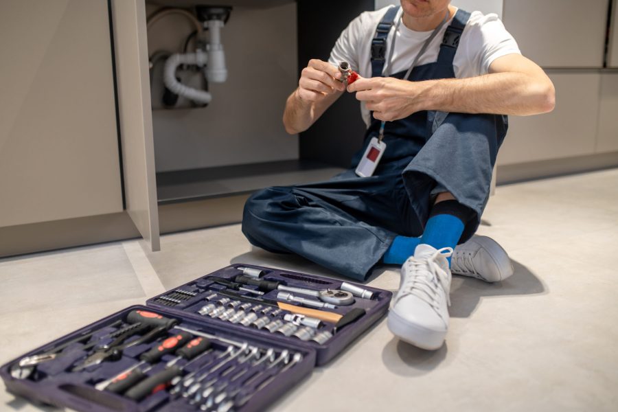 Man sitting on floor with plumbing detail in hands Right choice. Man in blue overalls looking at plumbing detail sitting on floor near open cabinet with sink and set of tools in kitchen, no face
